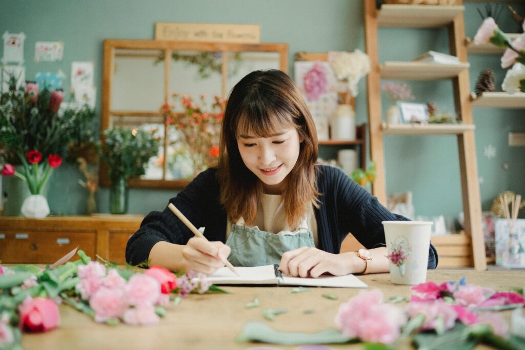 A young Asian woman florist writing notes in a vibrant floral workshop, smiling.